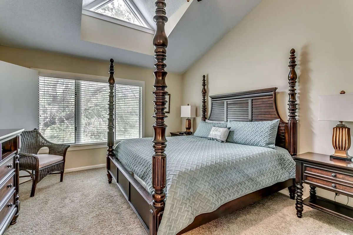 A serene primary bedroom featuring a four-poster king-sized bed, large windows that let in natural light, a woven chair in the corner, and a nightstand with a lamp. The room exudes a calming ambiance, perfect for relaxation and privacy.