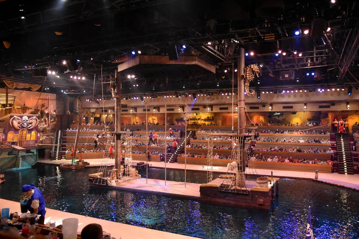 A vibrant interior scene showcasing a live show on a water-themed stage, with dramatic lighting, themed set design, and audience seating in the foreground. The energy of the performance and the engagement of the crowd are palpable.