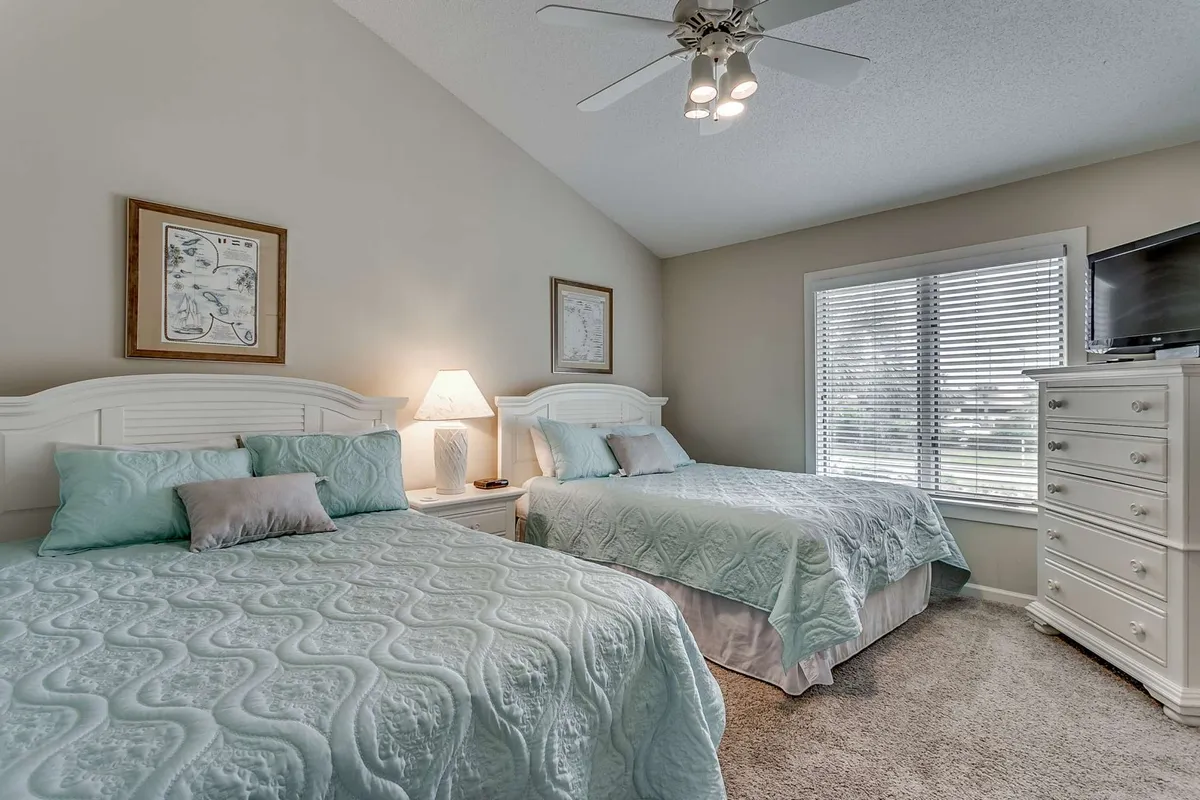 A cozy guest bedroom with two beds dressed in light blue bedding, a table lamp, framed artwork on the wall, a dresser topped with a TV, and sunny ambiance. The setup is ideal for families or friends seeking their own comfortable space.