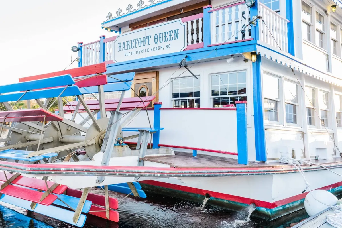 This vibrant exterior photo highlights the colorful Barefoot Queen paddleboat with its iconic paddle wheel, dock-side signage, and inviting atmosphere. Reflections in the windows and festive details evoke a sense of waterfront excitement and group enjoyment.