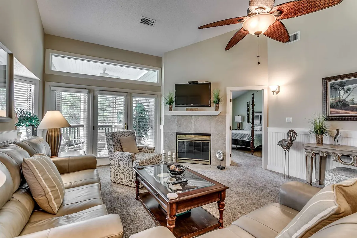 A spacious and inviting living room featuring a plush sofa, coffee table, fireplace, and television. Ample natural light streams in through large windows, creating a bright and cozy atmosphere perfect for relaxing or gathering with friends and family.